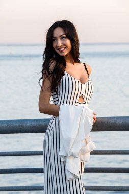 Çizgili siyah beyaz elbiseli güzel genç esmer kadın açık bir kadeh. Günbatımı sırasında iskelede duran kıvırcık kadın deniz arka plan bulanık 