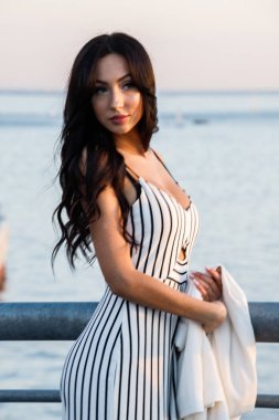Çizgili siyah beyaz elbiseli güzel genç esmer kadın açık bir kadeh. Günbatımı sırasında iskelede duran kıvırcık kadın deniz arka plan bulanık 
