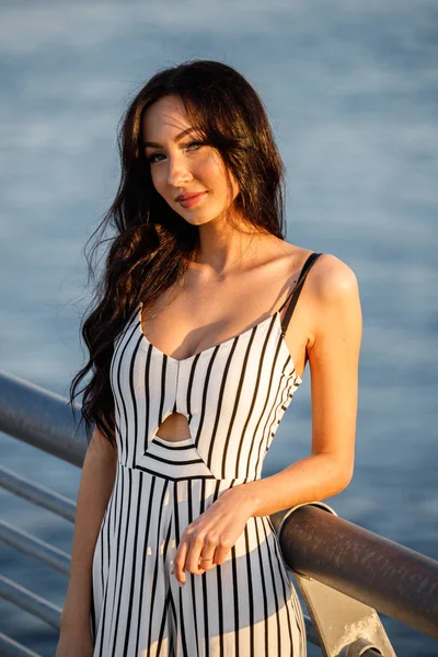 Çizgili siyah beyaz elbiseli güzel genç esmer kadın açık bir kadeh. Günbatımı sırasında iskelede duran kıvırcık kadın deniz arka plan bulanık 