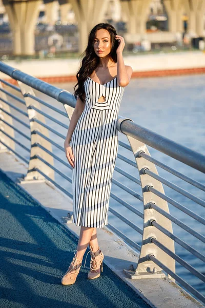 Çizgili siyah beyaz elbiseli güzel genç esmer kadın açık bir kadeh. Günbatımı sırasında iskelede duran kıvırcık kadın deniz arka plan bulanık 