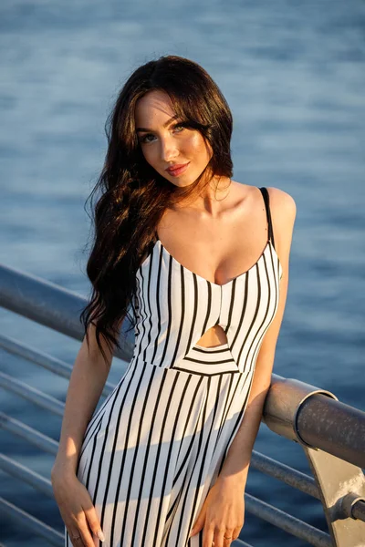 Çizgili siyah beyaz elbiseli güzel genç esmer kadın açık bir kadeh. Günbatımı sırasında iskelede duran kıvırcık kadın deniz arka plan bulanık 