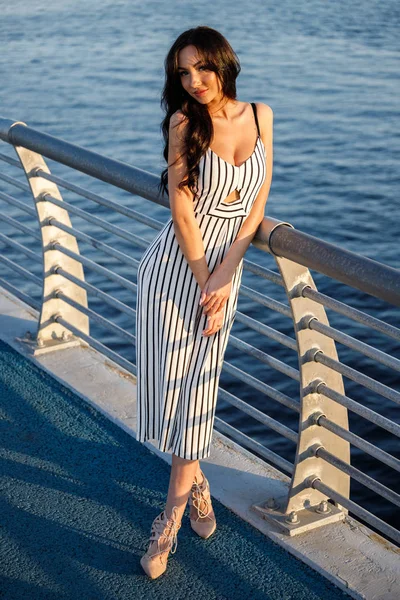 縞模様の白と黒のドレスで美しい若いブルネットの女性の屋外撮影 巻き毛の女性 日没時に桟橋に立って海の背景がぼやけてください — ストック写真