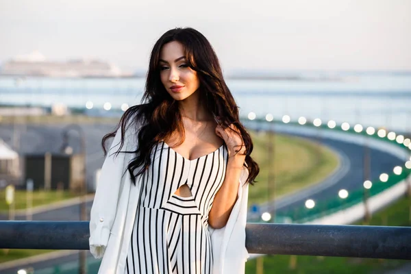Çizgili siyah beyaz elbiseli güzel genç esmer kadın açık bir kadeh. Güneş ışığı, şehir dolgu üzerinde duran kıvırcık kadın şehir görünümü arka plan bulanık 