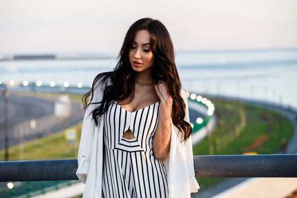 Çizgili siyah beyaz elbiseli güzel genç esmer kadın açık bir kadeh. Güneş ışığı, şehir dolgu üzerinde duran kıvırcık kadın şehir görünümü arka plan bulanık 