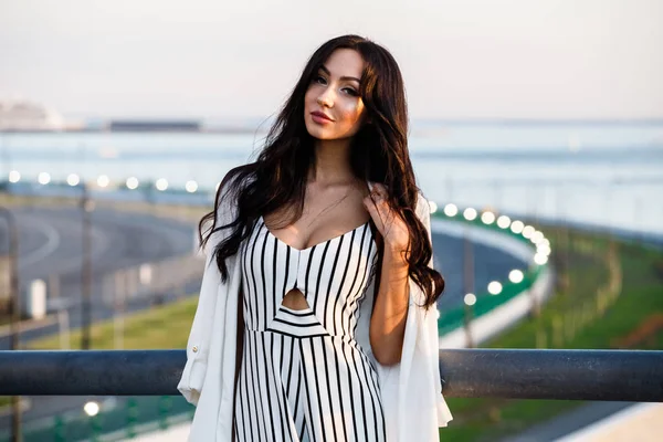 Çizgili siyah beyaz elbiseli güzel genç esmer kadın açık bir kadeh. Güneş ışığı, şehir dolgu üzerinde duran kıvırcık kadın şehir görünümü arka plan bulanık 