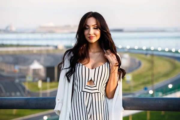 Çizgili siyah beyaz elbiseli güzel genç esmer kadın açık bir kadeh. Güneş ışığı, şehir dolgu üzerinde duran kıvırcık kadın şehir görünümü arka plan bulanık 