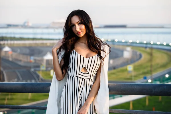 Çizgili siyah beyaz elbiseli güzel genç esmer kadın açık bir kadeh. Güneş ışığı, şehir dolgu üzerinde duran kıvırcık kadın şehir görünümü arka plan bulanık 