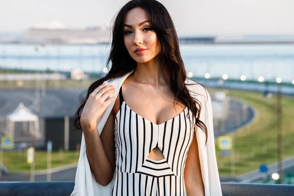 Çizgili siyah beyaz elbiseli güzel genç esmer kadın açık bir kadeh. Güneş ışığı, şehir dolgu üzerinde duran kıvırcık kadın şehir görünümü arka plan bulanık 