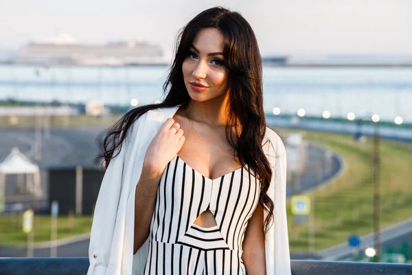 Çizgili siyah beyaz elbiseli güzel genç esmer kadın açık bir kadeh. Güneş ışığı, şehir dolgu üzerinde duran kıvırcık kadın şehir görünümü arka plan bulanık 