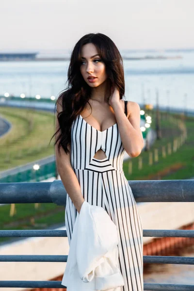 Çizgili siyah beyaz elbiseli güzel genç esmer kadın açık bir kadeh. Güneş ışığı, şehir dolgu üzerinde duran kıvırcık kadın şehir görünümü arka plan bulanık 