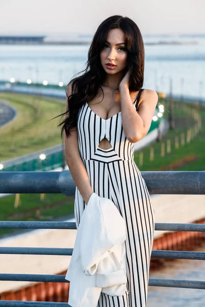 Çizgili siyah beyaz elbiseli güzel genç esmer kadın açık bir kadeh. Güneş ışığı, şehir dolgu üzerinde duran kıvırcık kadın şehir görünümü arka plan bulanık 
