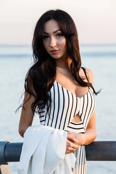 Çizgili siyah beyaz elbiseli güzel genç esmer kadın açık bir kadeh. Günbatımı sırasında iskelede duran kıvırcık kadın deniz arka plan bulanık 