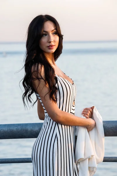 縞模様の白と黒のドレスで美しい若いブルネットの女性の屋外撮影 巻き毛の女性 日没時に桟橋に立って海の背景がぼやけてください — ストック写真
