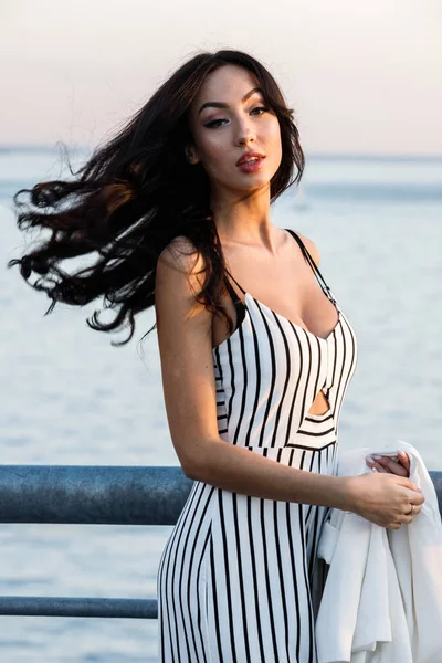 Çizgili siyah beyaz elbiseli güzel genç esmer kadın açık bir kadeh. Günbatımı sırasında iskelede duran kıvırcık kadın deniz arka plan bulanık 