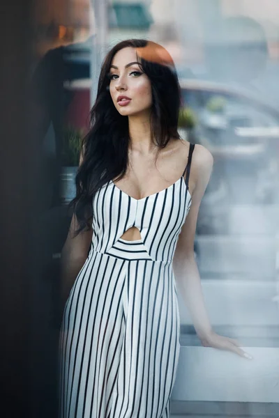 縞模様の白と黒のドレスで美しい若いブルネットの女性の屋外のポートレート カフェのインテリアに立っている巻き毛の女 — ストック写真