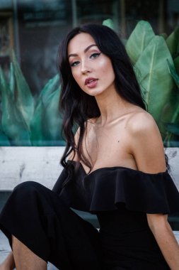 Yeşil bitkiler arka plan tahta bankta oturan şık makyaj ile siyah elbiseli güzel genç esmer kadın portresi