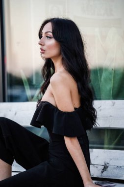 Yeşil bitkiler arka plan tahta bankta oturan şık makyaj ile siyah elbiseli güzel genç esmer kadın portresi