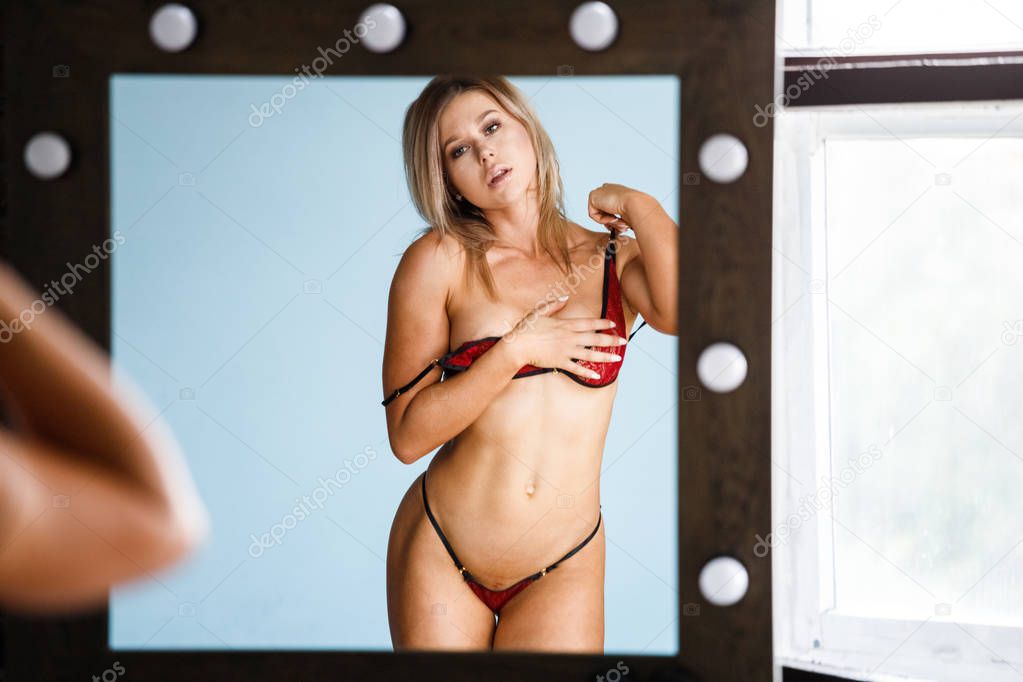 Studio portrait of beauty blond woman wearing red-black sexual lingerie standing close to mirror on blue wall background 