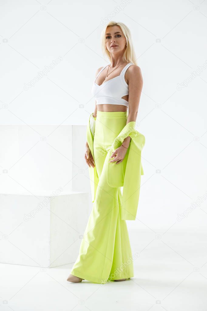 Fashion studio portrait of beauty blond model in white room interior. Shot of beauty young woman wearing green casual suit and white shirt