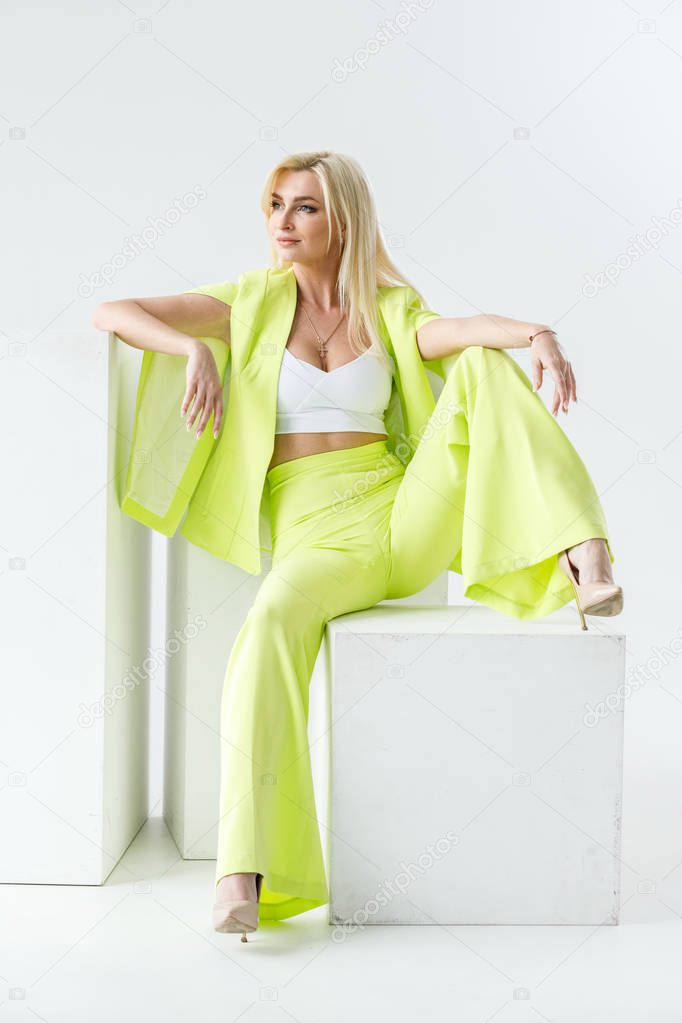 Fashion studio portrait of beauty blond model in white room interior. Shot of beauty young woman wearing green casual suit and white shirt