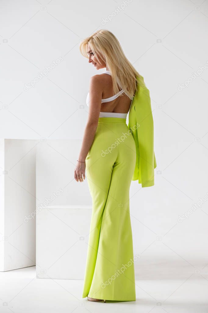 Fashion studio portrait of beauty blond model in white room interior. Shot of beauty young woman wearing green casual suit and white shirt