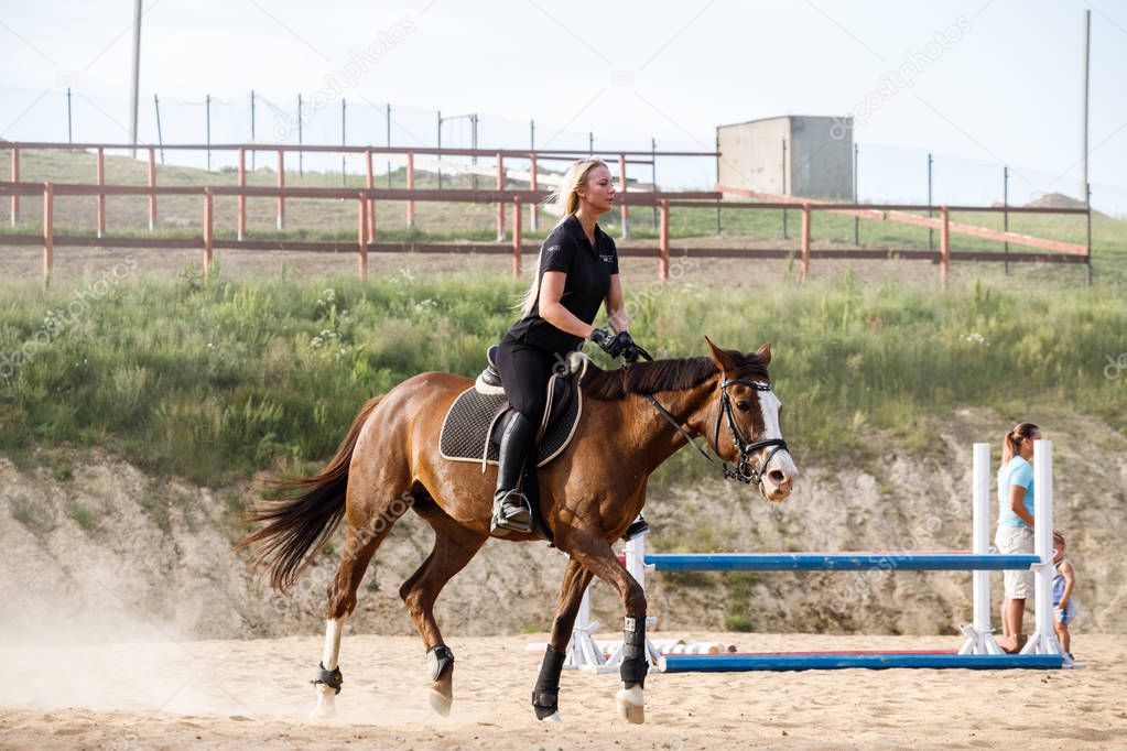 Outdoor shot of beauty blond woman riding horse at daytime. Young woman in sporty clothes enjoying horseback riding at racecourse 