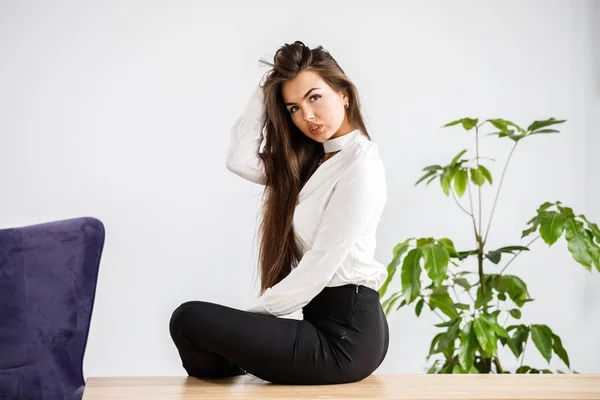 白いシャツと黒のズボンで若いブルネットの女性の肖像画スタジオ オフィスのインテリアにテーブルの上に座って美容ビジネス女性 — ストック写真