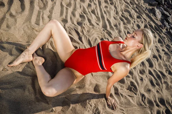 美しい金髪の女性とスポーティなボディが着ている赤い水着の肖像画 昼で砂浜に座って美しいモデル — ストック写真