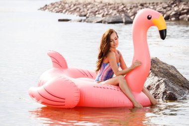 Güzellik esmer renkli yaz mayo modelinde açık portresi. Gündüz, dalgalı su arka plan Flamingo'da şeklinde pembe şişme yatak üzerinde oturan genç güzel kadın