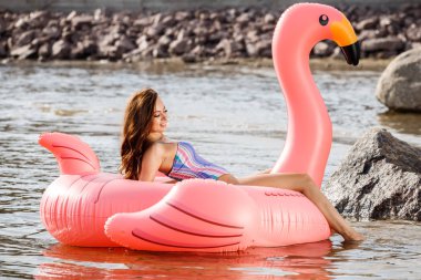 Güzellik esmer renkli yaz mayo modelinde açık portresi. Gündüz, dalgalı su arka plan Flamingo'da şeklinde pembe şişme yatak üzerinde oturan genç güzel kadın