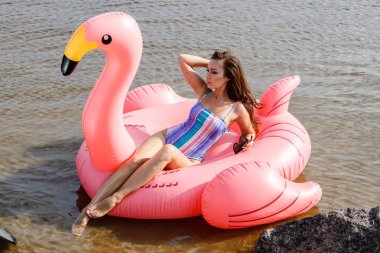 Güzellik esmer renkli yaz mayo modelinde açık portresi. Gündüz, dalgalı su arka plan Flamingo'da şeklinde pembe şişme yatak üzerinde oturan genç güzel kadın