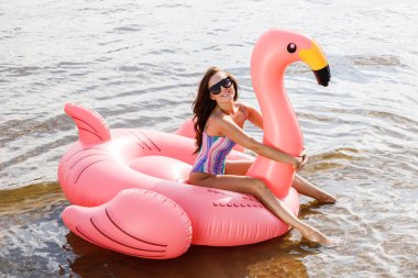 Güzellik esmer renkli yaz mayo modelinde açık portresi. Gündüz, dalgalı su arka plan Flamingo'da şeklinde pembe şişme yatak üzerinde oturan genç güzel kadın