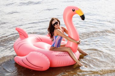 Güzellik esmer renkli yaz mayo modelinde açık portresi. Gündüz, dalgalı su arka plan Flamingo'da şeklinde pembe şişme yatak üzerinde oturan genç güzel kadın