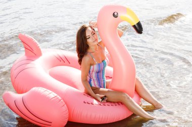 Güzellik esmer renkli yaz mayo modelinde açık portresi. Gündüz, dalgalı su arka plan Flamingo'da şeklinde pembe şişme yatak üzerinde oturan genç güzel kadın