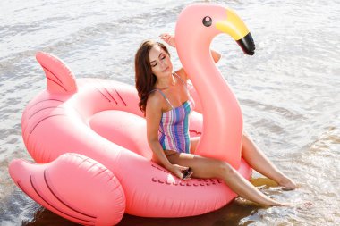 Güzellik esmer renkli yaz mayo modelinde açık portresi. Gündüz, dalgalı su arka plan Flamingo'da şeklinde pembe şişme yatak üzerinde oturan genç güzel kadın