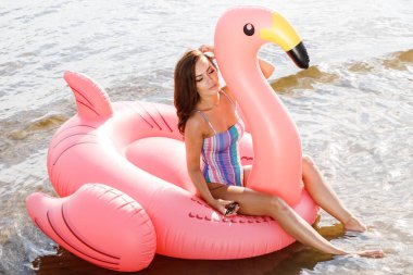 Güzellik esmer renkli yaz mayo modelinde açık portresi. Gündüz, dalgalı su arka plan Flamingo'da şeklinde pembe şişme yatak üzerinde oturan genç güzel kadın