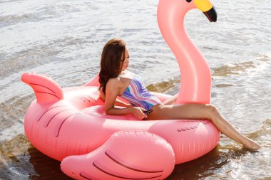 Güzellik esmer renkli yaz mayo modelinde açık portresi. Gündüz, dalgalı su arka plan Flamingo'da şeklinde pembe şişme yatak üzerinde oturan genç güzel kadın