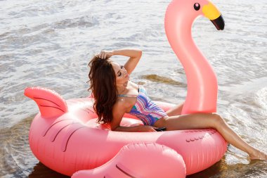 Güzellik esmer renkli yaz mayo modelinde açık portresi. Gündüz, dalgalı su arka plan Flamingo'da şeklinde pembe şişme yatak üzerinde oturan genç güzel kadın