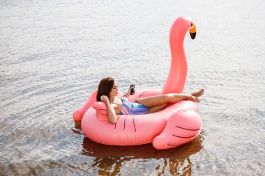 Güzellik esmer renkli yaz mayo modelinde açık portresi. Gündüz, dalgalı su arka plan Flamingo'da şeklinde pembe şişme yatak üzerinde oturan genç güzel kadın