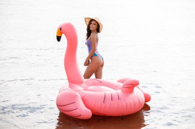 Güzellik esmer renkli yaz mayo modelinde açık portresi. Gündüz, dalgalı su arka plan Flamingo'da şeklinde pembe şişme yatak üzerinde oturan genç güzel kadın