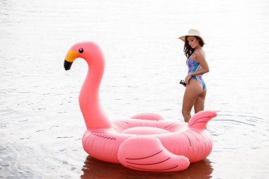 Güzellik esmer renkli yaz mayo modelinde açık portresi. Gündüz, dalgalı su arka plan Flamingo'da şeklinde pembe şişme yatak üzerinde oturan genç güzel kadın
