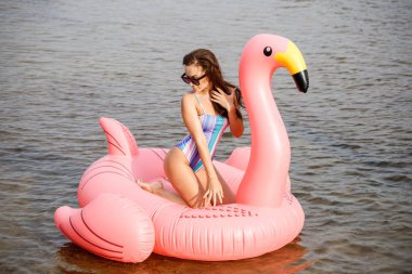 Güzellik esmer renkli yaz mayo modelinde açık portresi. Gündüz, dalgalı su arka plan Flamingo'da şeklinde pembe şişme yatak üzerinde oturan genç güzel kadın