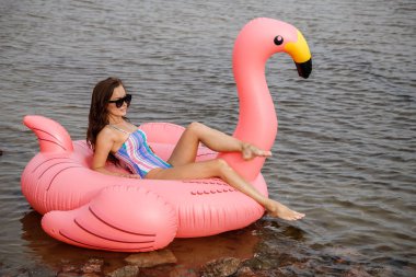 Güzellik esmer renkli yaz mayo modelinde açık portresi. Gündüz, dalgalı su arka plan Flamingo'da şeklinde pembe şişme yatak üzerinde oturan genç güzel kadın