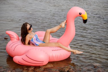 Güzellik esmer renkli yaz mayo modelinde açık portresi. Gündüz, dalgalı su arka plan Flamingo'da şeklinde pembe şişme yatak üzerinde oturan genç güzel kadın