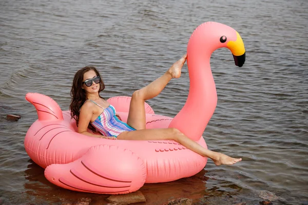 カラフルな夏の水着で美ブルネット モデルの屋外のポートレート 波状水背景でフラミンゴの形でピンクの膨脹可能なマットレスの上に座っていた美しい女性 — ストック写真
