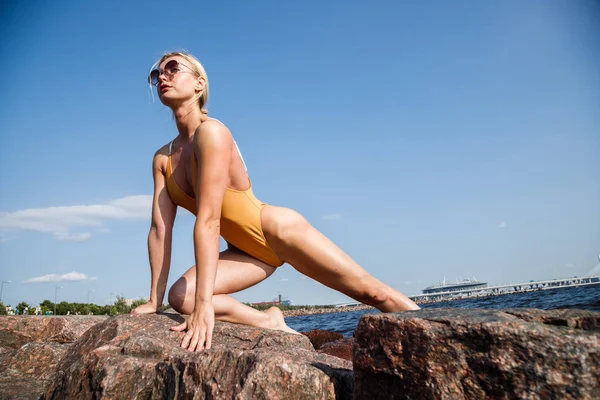 白天戴着太阳镜和泳衣的金发美女在岩石海滩上健身 — 图库照片
