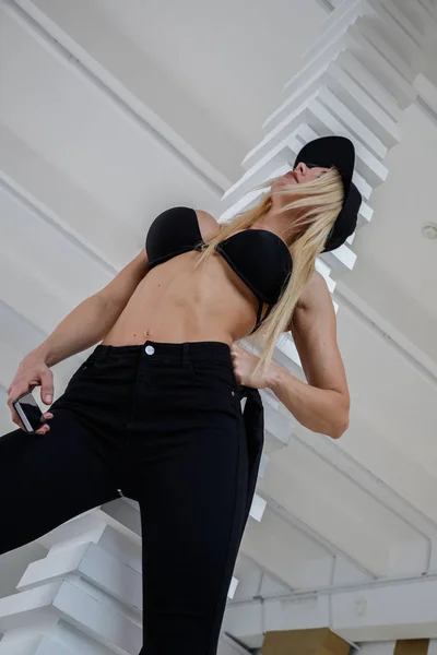 高級アパートのインテリアでスポーティな金髪モデルのスタジオ撮影 若い女性の手の中のスマート フォンで立っている黒い服を着て — ストック写真