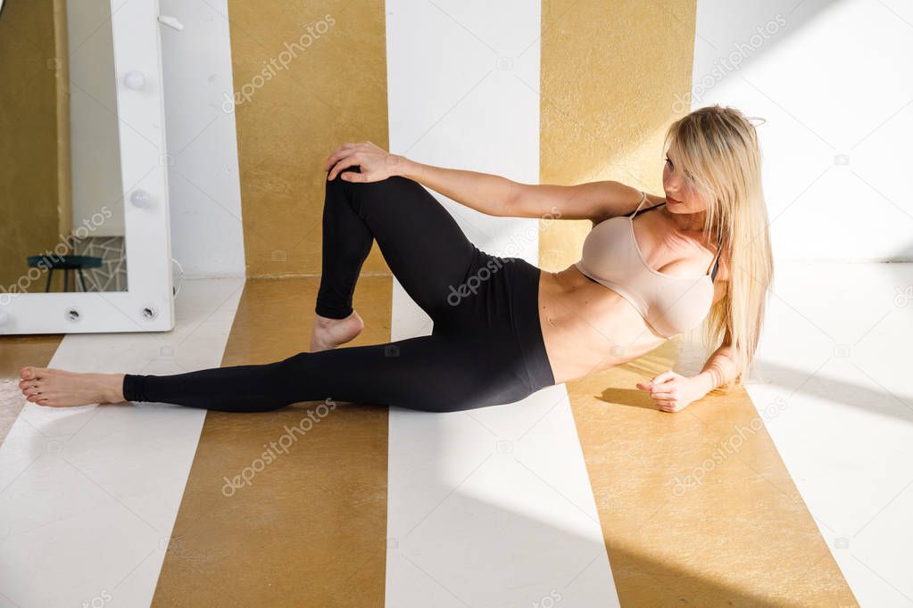 Studio shot of sporty blond model in luxury apartments interior. Young woman wearing black leggings and beige top lying on floor at hotel interior 