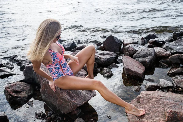 白天穿着太阳镜和泳衣站在岩石海滩上的金发美女 — 图库照片