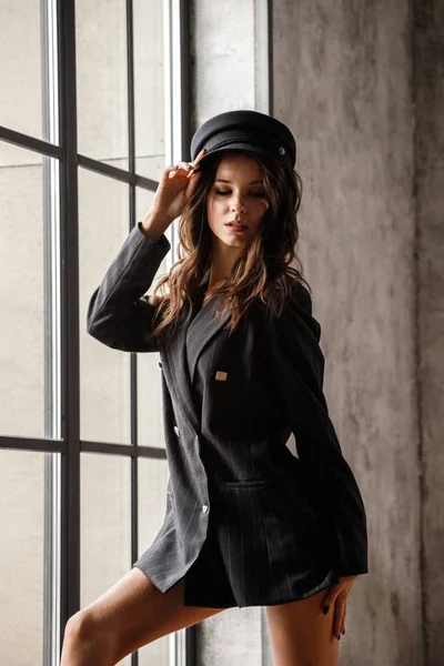 窓近くに美容女性モデル立ちの肖像画 モダンなインテリアのグレーのジャケットと灰色の帽子を着ているブルネットの女性 — ストック写真